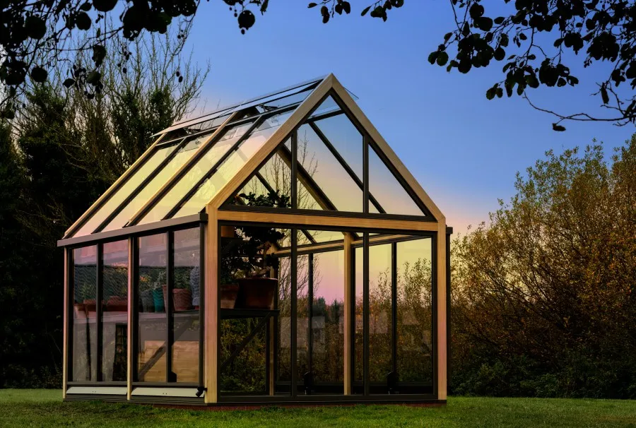 A modern, glass greenhouse with a wooden frame, standing on a grassy lawn at dusk.